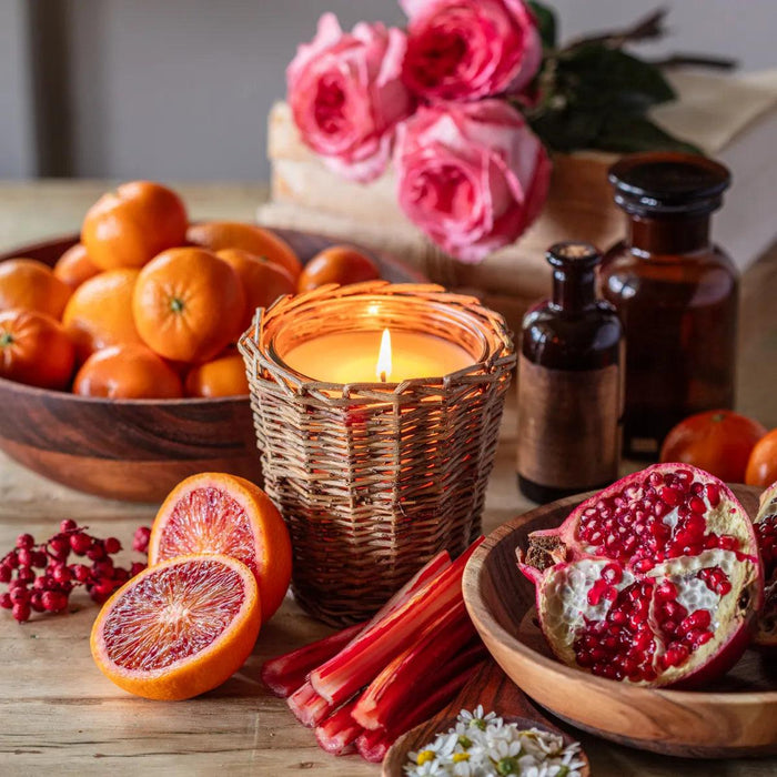 Lit candle in a woven wicker holder surrounded by fresh red currants, blood oranges, and pomegranate on a wooden table.