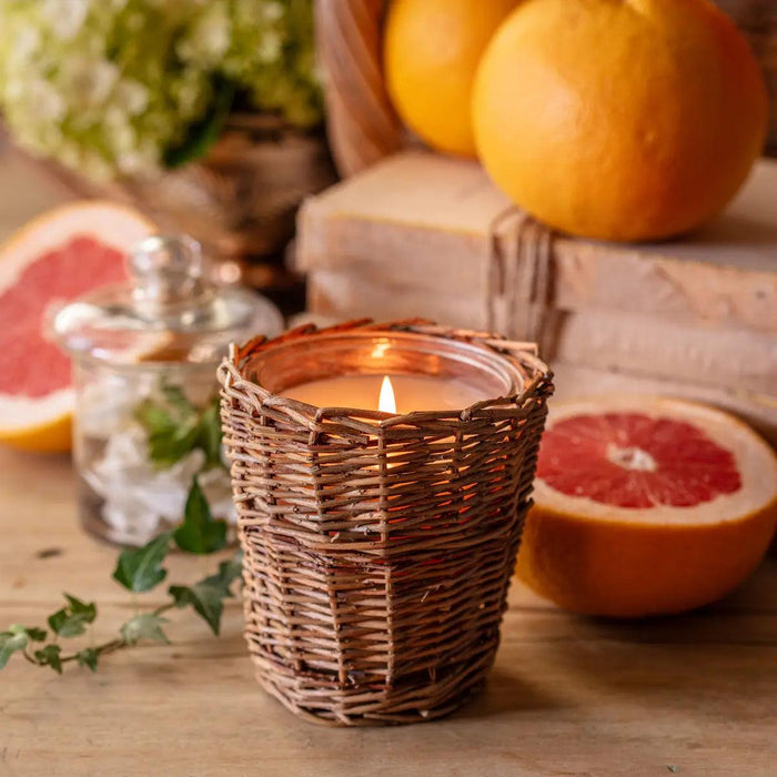 Lit candle in a rustic woven wicker holder surrounded by fresh grapefruit halves and greenery on a wooden table.