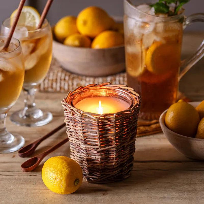Lit candle in a woven willow basket holder surrounded by fresh lemons and glasses of iced tea on a wooden table.