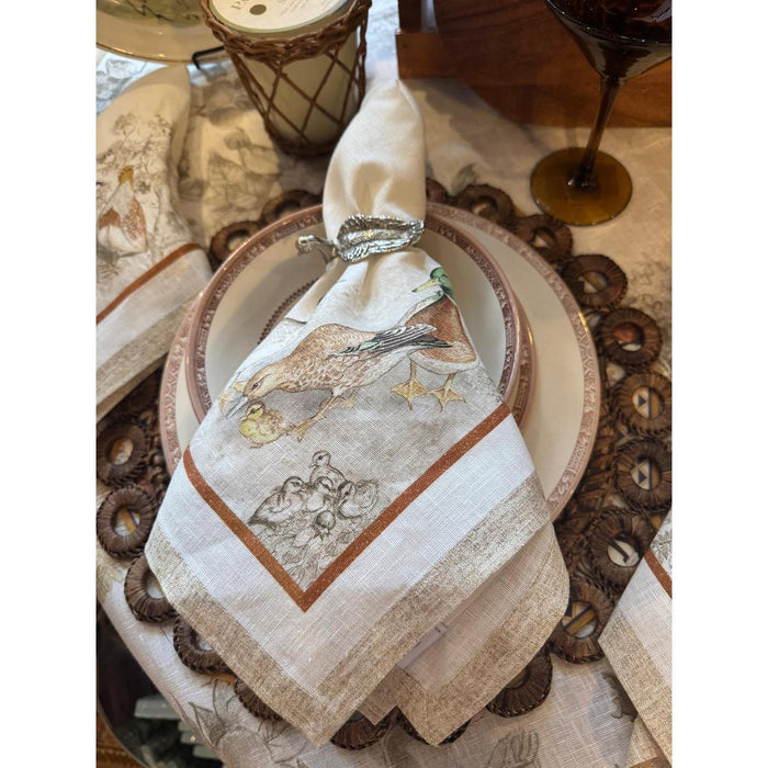 Cream-colored Italian linen napkin featuring detailed gamebird illustrations and a silver leaf napkin ring on a place setting.