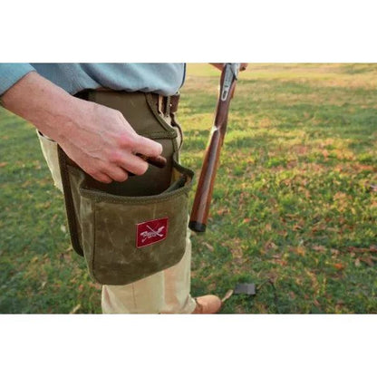Olive green waxed canvas shotshell belt pouch with a red logo patch, worn on a belt while holding a shotgun.