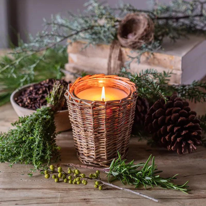 Lit candle in a woven willow holder surrounded by pine needles, herbs, and a pinecone on a rustic wooden table.