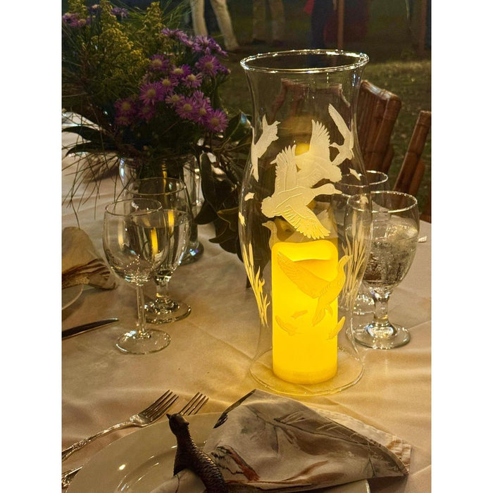 Clear glass hurricane centerpiece hand-etched with flying quail and waterfowl, glowing with a warm LED pillar candle.