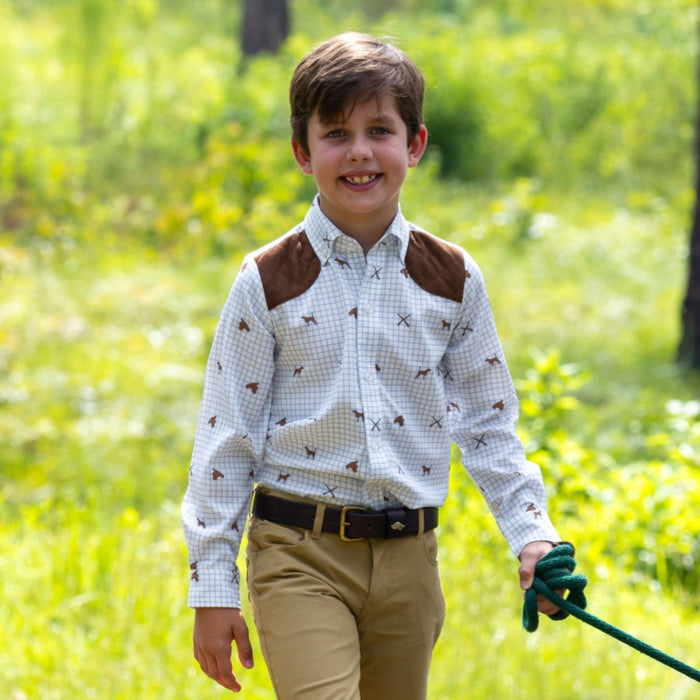Boy wearing a white and blue checkered long-sleeve performance shooting shirt with brown shoulder patches outdoors.