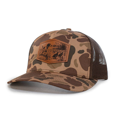 Camo Richardson 112 trucker cap with a brown leather patch featuring an embossed quail and pointer dog scene.