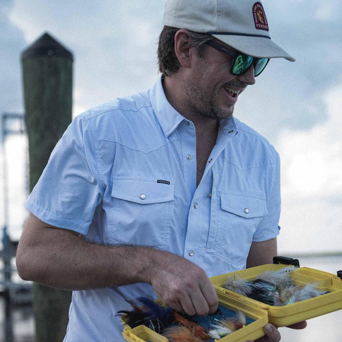 Man wearing a light blue short-sleeve western shirt with pearl snaps and chest pockets while holding a fishing tackle box.