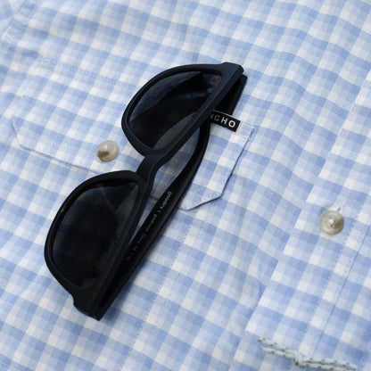 Black sunglasses resting in the chest pocket of a light blue and white gingham Poncho performance shirt.