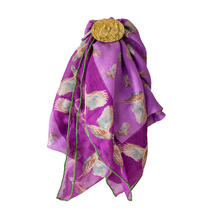 Brass quail trio scarf ring securing a purple patterned scarf with bird illustrations and green stitched edges.