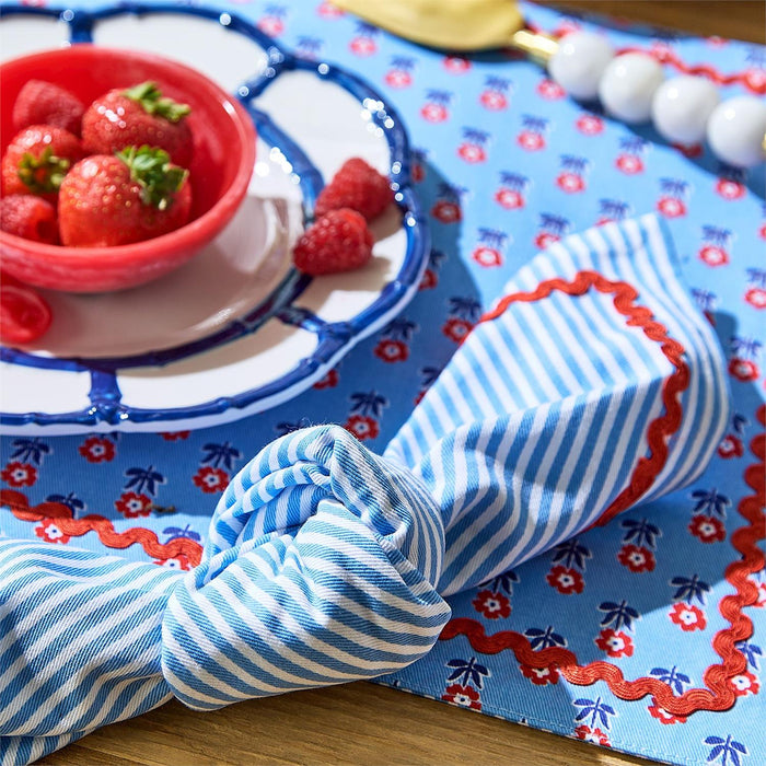 Cotton dinner napkin with blue stripes and red ric rac trim, styled on a matching floral placemat with a bowl of strawberries.