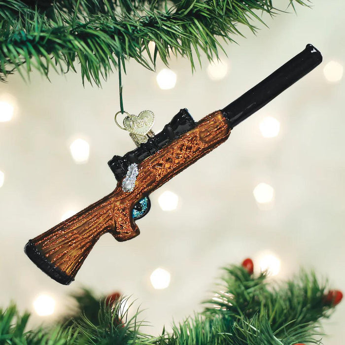 Hand-painted blown glass rifle ornament with glittered brown wood-grain stock and black barrel hanging on a tree.