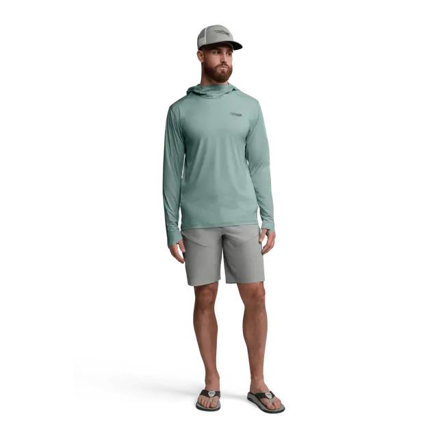 Man wearing a sage green Sitka Guard Sun Hoodie with a hood, thumb loops, and grey shorts against a white background.
