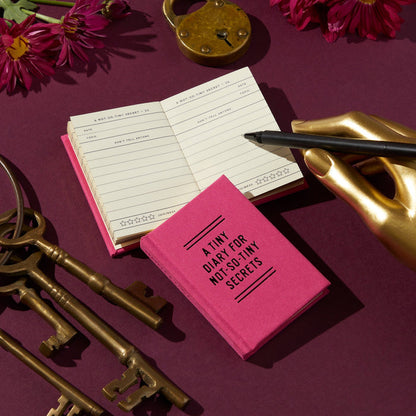 A Tiny Diary for Not So Tiny Secrets by Brass Monkey, in pink with gold-edged pages, lies open on a burgundy surface. Nearby are keys, a lock, flowers, and a pen held upright in a gold hand-shaped holder.