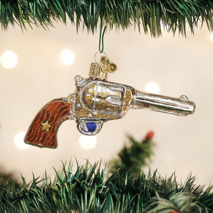 Hand-painted blown glass revolver ornament with gold glitter accents and a wood-grain handle hanging on a Christmas tree.